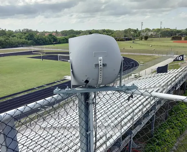 Outdoor security camera antenna mounted on a metal fence overlooking a green landscape under a clear sky.