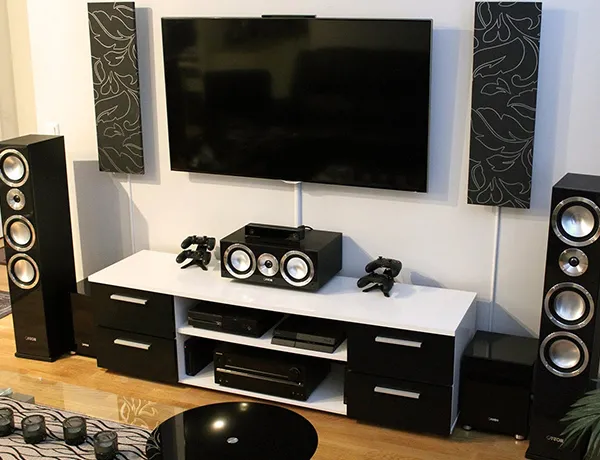 Home theater setup with flat screen TV mounted above a black and white media console, surrounded by tall floor-standing speakers and a central speaker in a modern living room.