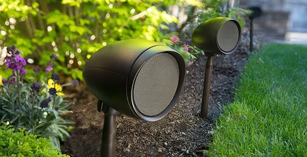 Black cylindrical outdoor speakers installed in garden landscaping with grass and plants, illustrating outdoor speaker setup in Boca Raton, FL.